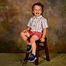 Rhettlee joined the competition — help win amazing prizes! child, smiling, curly_hair, striped_shirt, shorts, sneakers, dinosaur_design, wooden_stool, studio_background, portrait, happy, sitting, indoor, toddler, fashion, clothing, feet, face, person, photography