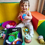 Abygaïl a rejoint le concours — aidez-le/la à gagner de superbes lots ! baby, child, smiling, toys, plastic_discs, colorful, play_area, padded_furniture, purple_bowl, basket, floor, clothing, socks, happy, indoors, person, face, head, playtime, cute