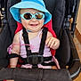 baby, stroller, sunglasses, hat, smile, child, seatbelt, pink_clothing, sunhat, outdoor, happy, infant, cute, baby_toy, black_stroller, safety_harness, person, skin, legs, hand