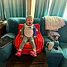 toddler, child, red_chair, car_shaped_chair, living_room, couch, curtains, laptop, remote_control, purse, drink_cup, blanket, socks, sneakers, wooden_beam, carpet, person, indoor, expression, furniture