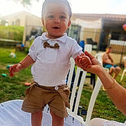 Kinay a rejoint le concours — aidez-le/la à gagner de superbes lots ! toddler, child, smiling, standing, holding_hand, white_shirt, bow_tie, brown_shorts, socks, outdoor, grass, chair, adult_hand, sunlight, backyard, person, casual, happy, daytime, portrait
