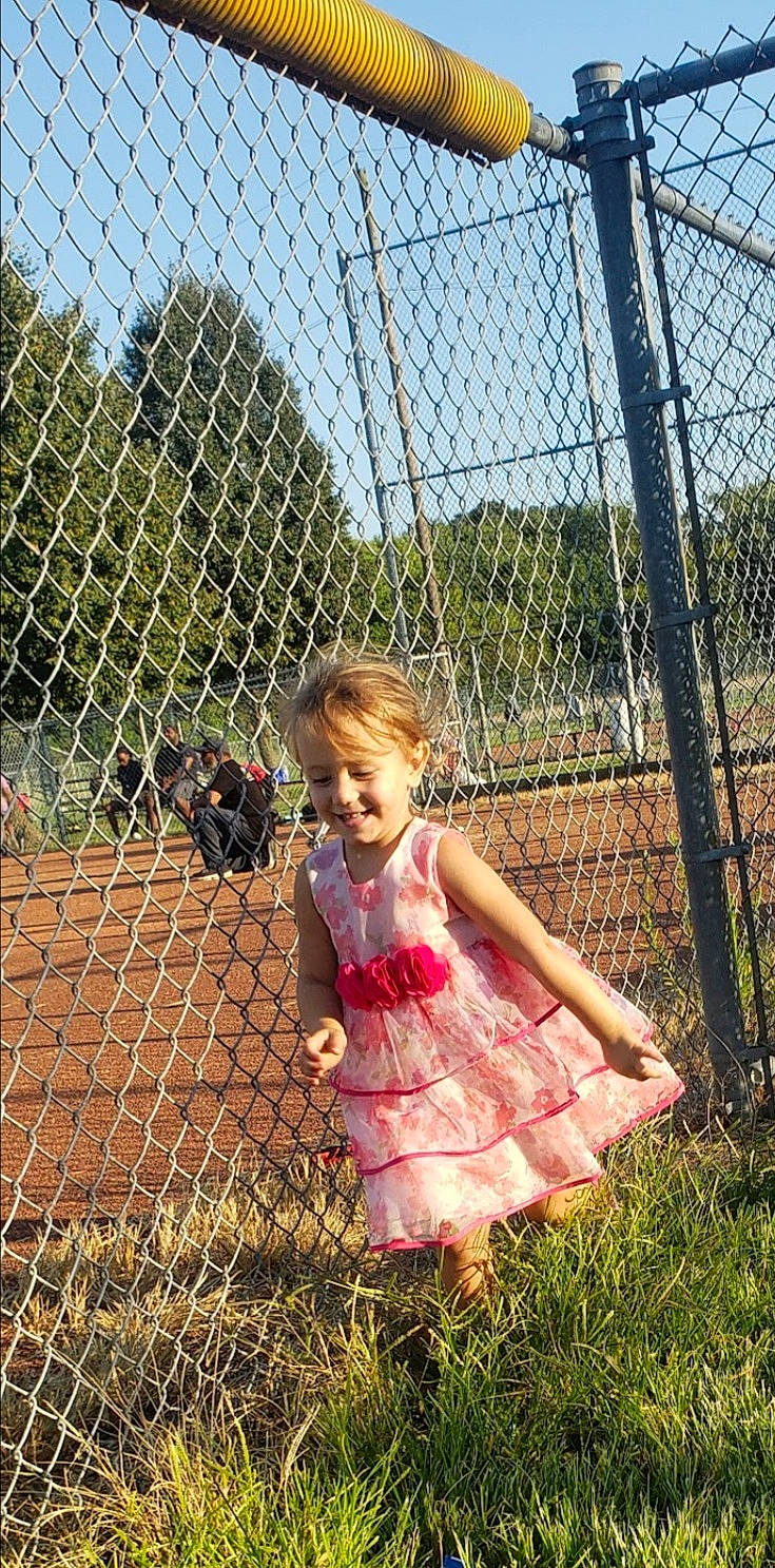 Krystol is registered to the contest to win money with this photo: baby_toddler_clothing, chain_link_fencing, dress, fence, fun, grass, happy, joy, leisure, mammal, mesh, nature, people_in_nature, person, plant, sky, smile, toddler, tree, vertebrate