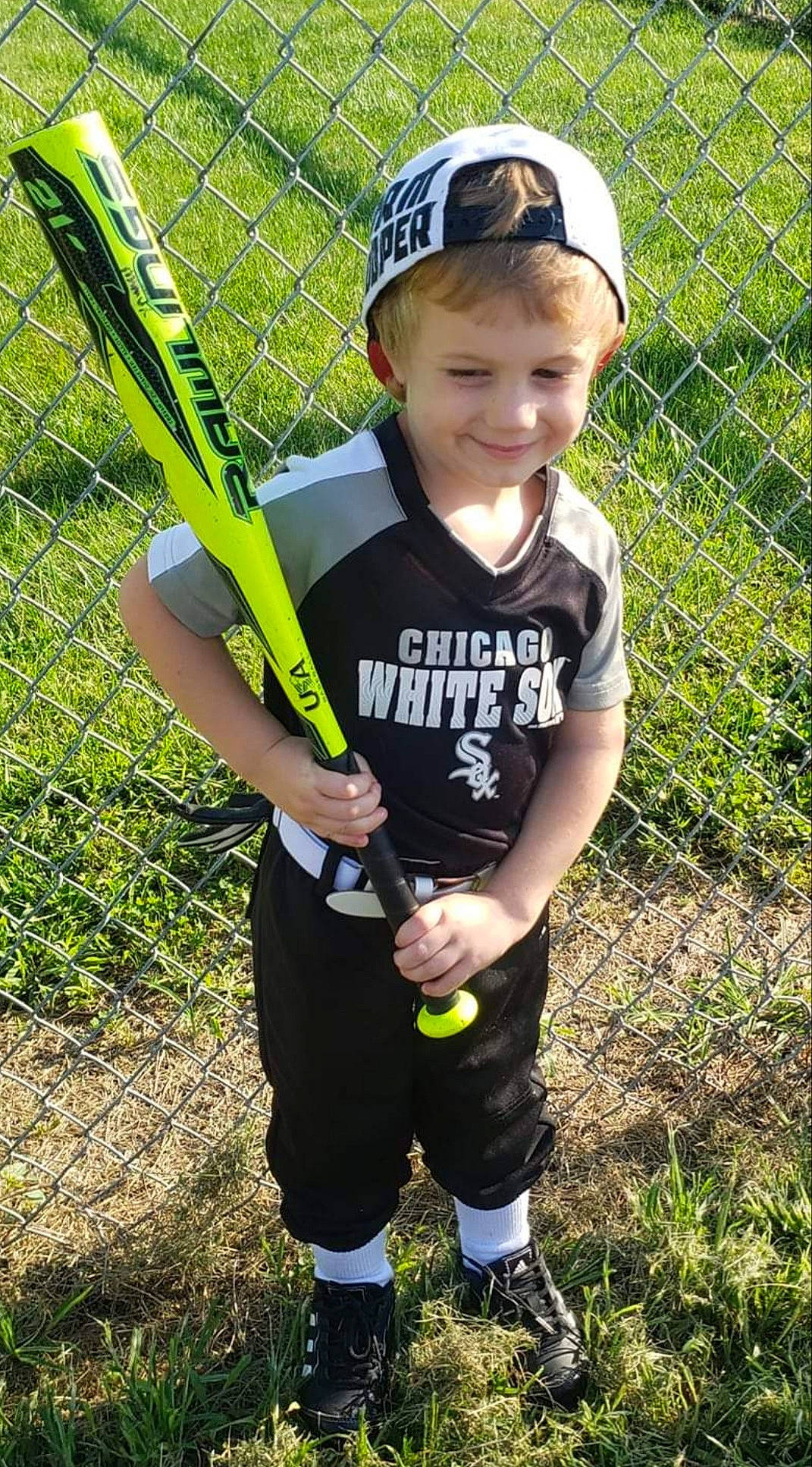 Kayden is registered to the contest to win money with this photo: baseball_bat, baseball_cap, baseball_equipment, cap, child, fence, fun, goggles, grass, joy, leisure, mesh, people_in_nature, person, personal_protective_equipment, shorts, smile, soil, sports_equipment, t_shirt