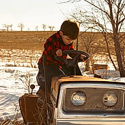 Archer is registered to the contest to win money with this photo: child, boy, lawn_tractor, snow, winter, outdoor, tree, field, rusty, steering_wheel, red_shirt, jeans, nature, daylight, rural, cold, curious, playing, vehicle, season