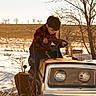 child, boy, lawn_tractor, snow, winter, outdoor, tree, field, rusty, steering_wheel, red_shirt, jeans, nature, daylight, rural, cold, curious, playing, vehicle, season