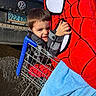 child, shopping_cart, plush_toy, spiderman, towel, car, license_plate, outdoor, happy, smiling, clothing, toy, person, kid, blue, red, metal, vehicle, daylight, parking_lot