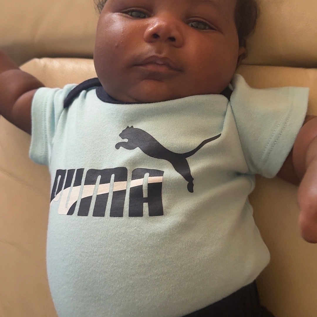 Delijah is registered to the contest to win money with this photo: baby, child, clothing, comfort, couch, curly_hair, expression, face, furniture, indoor, infant, pants, person, portrait, puma, relaxed, shirt, sitting, skin, young