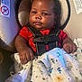 baby, infant, car_seat, blanket, red_clothing, child, seat_belt, curly_hair, indoors, cute, chubby_cheeks, holding, fabric, patterned_blanket, safety, portrait, young_child, closeup, infant_baby, baby_hands