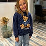 blonde_hair, casual_clothing, chair, child, desk, dried_flowers, flooring, girl, happy, home, indoor, jeans, portrait, room, smiling, standing, sweatshirt, teddy_bear, vase, young