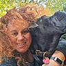 woman, dog, pet, smile, outdoor, curly_hair, leash, collar, affection, close_up, portrait, natural_light, watch, red_hair, heart_shaped_tag, friendship, greenery, happy, casual_clothing, animal