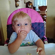 Mason is registered to the contest to win money with this photo: toddler, child, high_chair, snack, blue_eyes, blond_hair, indoor, furniture, curtain, table, hand, face, person, clothing, seat, home, eating, cute, portrait, young_child