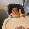 baby, high_chair, bib, yellow_bib, curious, indoor, hand, rings, cleaning, food_smear, wall, appliance, face, child, person, clothing, table, feeding, infant, expression