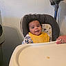 baby, child, high_chair, yellow_bib, curly_hair, smile, face, hand, rings, tray, vacuum_cleaner, wall, indoor, person, clothing, furniture, happy, seated, infant, cute