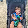 Ayden is registered to the contest to win money with this photo: toddler, stroller, child, water_bottle, blue_clothing, outdoor, gravel_path, people, fence, dinosaur_design, curious_expression, sitting, baby, person, summer, daylight, casual, hand, legs, face