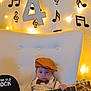 baby, child, guitar, music_notes, fairy_lights, chair, brown_cap, bow_tie, white_shirt, suspenders, tufted_chair, musical_theme, indoors, decor, portrait, cute, young_child, string_instrument, lighting, cozy
