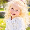 blonde_hair, blue_eyes, child, curly_hair, cute, daylight, face, girl, grass, happy, nature, outdoor, park, person, pink_bow, portrait, smiling, sunlight, tree, white_jacket