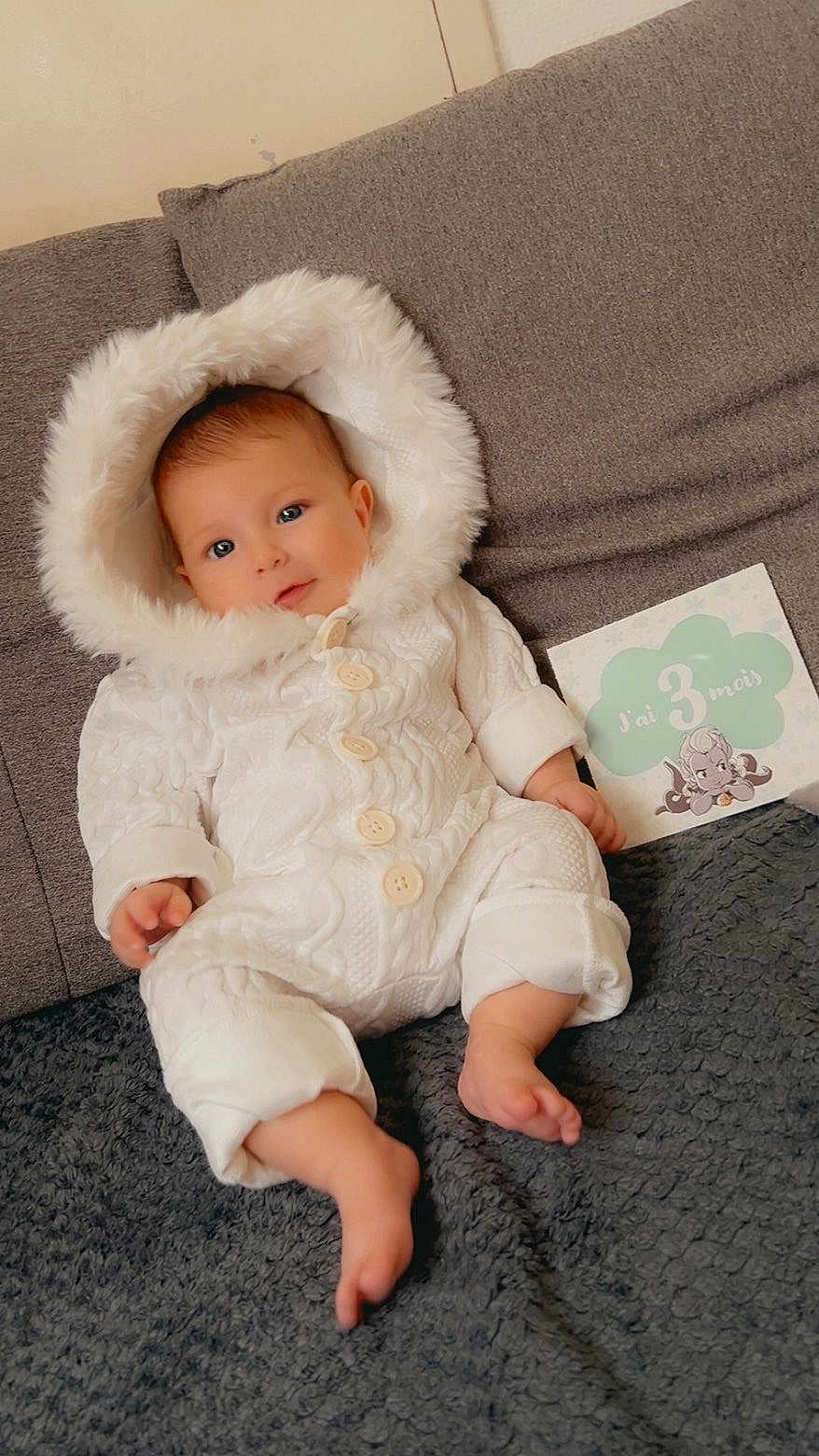 Amaya participe au concours pour gagner de l'argent avec cette photo : baby, infant, white_onesie, hood, couch, blanket, milestone_card, 3_months, cute, cozy, indoors, smiling, portrait, child, person, soft_texture, warm_clothing, happy, sitting, face