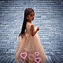 child, girl, dress, peach_dress, long_hair, brick_wall, wood_floor, hearts, pink_hearts, back_view, looking_back, indoor, portrait, flooring, wall, cute, young, standing, soft_lighting, whimsical