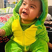 Noah joined the competition — help win amazing prizes! baby, child, dinosaur_costume, green, yellow, smiling, happy, cute, plush, hood, indoors, bed, blanket, person, infant, sitting, cozy, soft, clothing, portrait