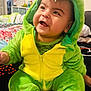 baby, child, dinosaur_costume, green, yellow, smiling, happy, cute, plush, hood, indoors, bed, blanket, person, infant, sitting, cozy, soft, clothing, portrait
