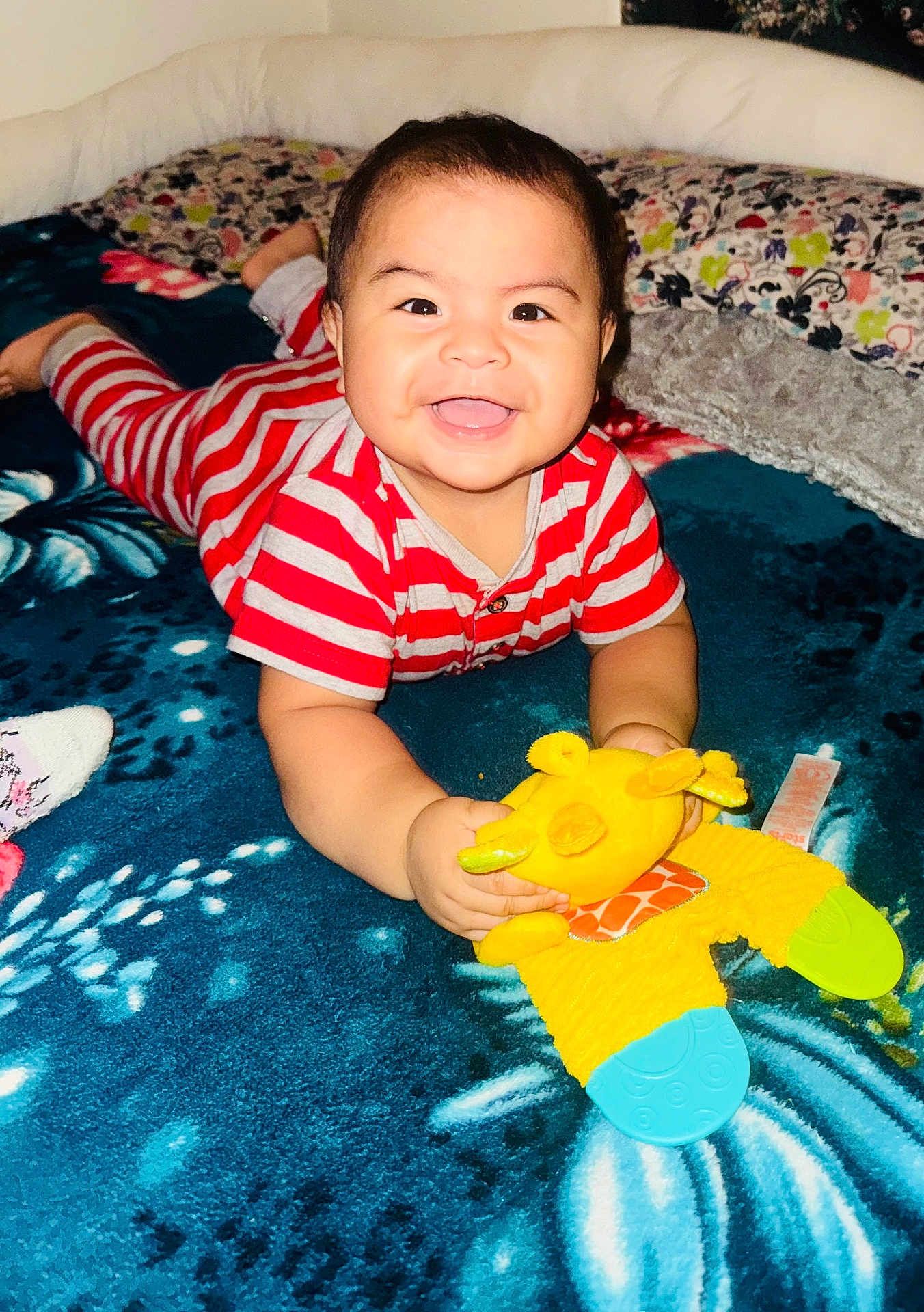 Noah is registered to the contest to win money with this photo: baby, smiling, toy, plush_toy, striped_clothing, blue_blanket, indoor, happy, child, cute, lying_down, teething_toy, face, bright_colors, cozy, infant, playful, young_child, cheerful, closeup