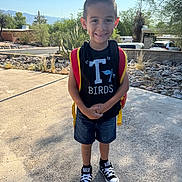 Antonio is registered to the contest to win money with this photo: backpack, boy, cactus, child, converse, daylight, desert_plants, outdoor, portrait, residential_area, road, shorts, sidewalk, sky, smiling, sneakers, standing, sunny, t_shirt, trees