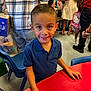 Antonio joined the competition — help win amazing prizes! backpack, blue_shirt, boy, casual_clothing, chair, child, classroom, education, face, floor, hair, hello_kitty, indoor, keys, people, person, red_table, smiling, table, toy_guitar_keychain
