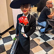 Rose Tuccino is registered to the contest to win money with this photo: child, mariachi, sombrero, traditional_clothing, red_bow, musical_instrument_case, smiling, indoor, checkered_floor, person, man, glasses, seated, black_jacket, white_shirt, black_skirt, earrings, floor_tiles, table, chair