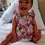 Renesmée joined the competition — help win amazing prizes! baby, child, smiling, sitting, bed, pillow, floral_dress, happy, infant, indoor, cute, face, headboard, stuffed_animal, toy, room, skin, person, young, furniture