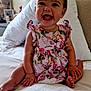 baby, child, smiling, sitting, bed, pillow, floral_dress, happy, infant, indoor, cute, face, headboard, stuffed_animal, toy, room, skin, person, young, furniture
