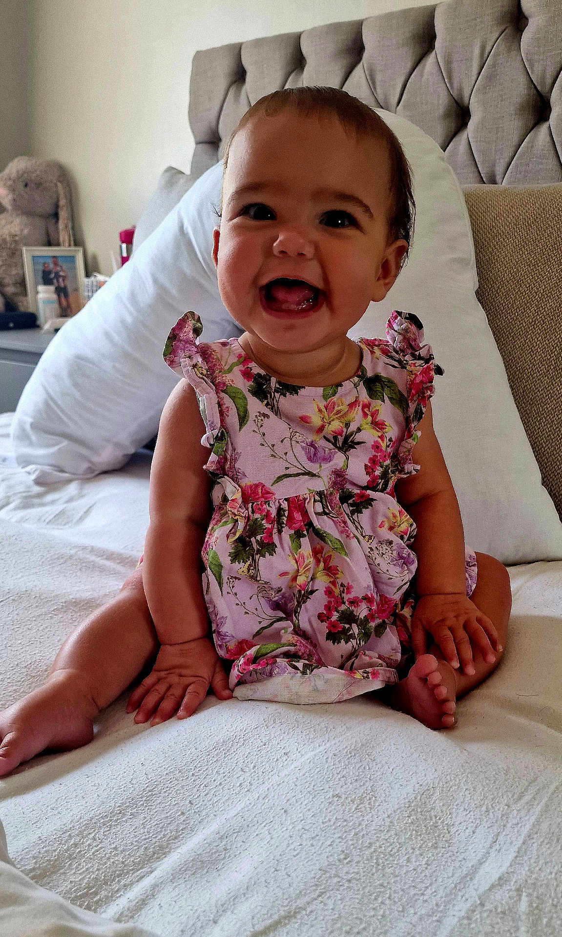 Renesmée joined the competition — help win amazing prizes! baby, child, smiling, sitting, bed, pillow, floral_dress, happy, infant, indoor, cute, face, headboard, stuffed_animal, toy, room, skin, person, young, furniture