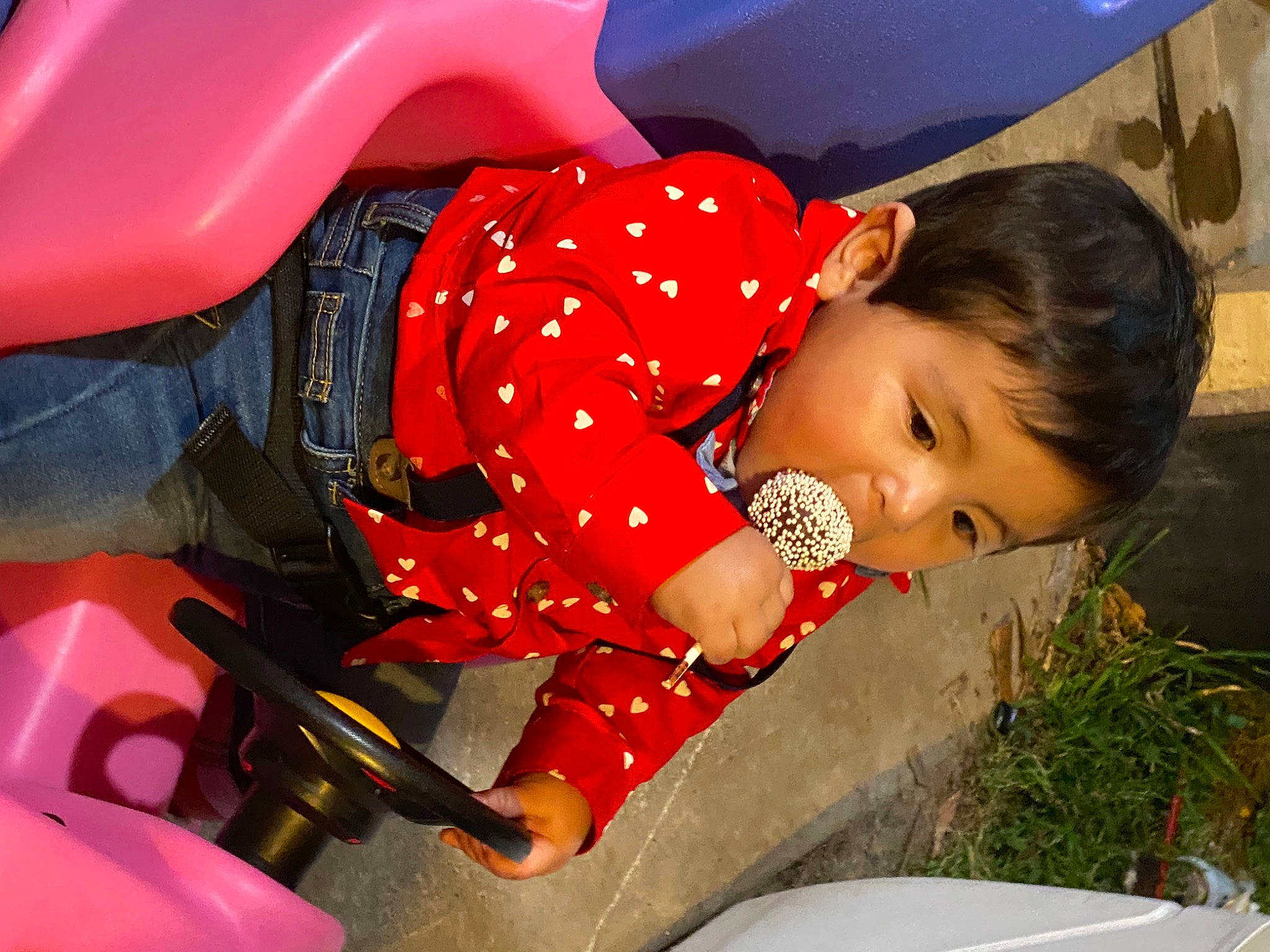 Cristian is registered to the contest to win money with this photo: black_hair, child, dress, face, fun, grass, hairstyle, human, human_leg, leisure, motor_vehicle, mouth, pattern, person, pink, plant, red, sitting, skin, thigh