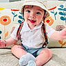 baby, child, smiling, hat, striped, bunny_ears, suspenders, white_shirt, blue_shorts, socks, couch, cushions, floral_pattern, indoor, cute, happy, portrait, sitting, toddler, cozy