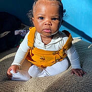 Ryleigh is registered to the contest to win money with this photo: toddler, child, baby, sitting, yellow_overalls, white_shirt, hair_accessories, curly_hair, indoors, blue_wall, blanket, sunlight, shadow, expression, portrait, cozy, clothing, person, cute, young_child