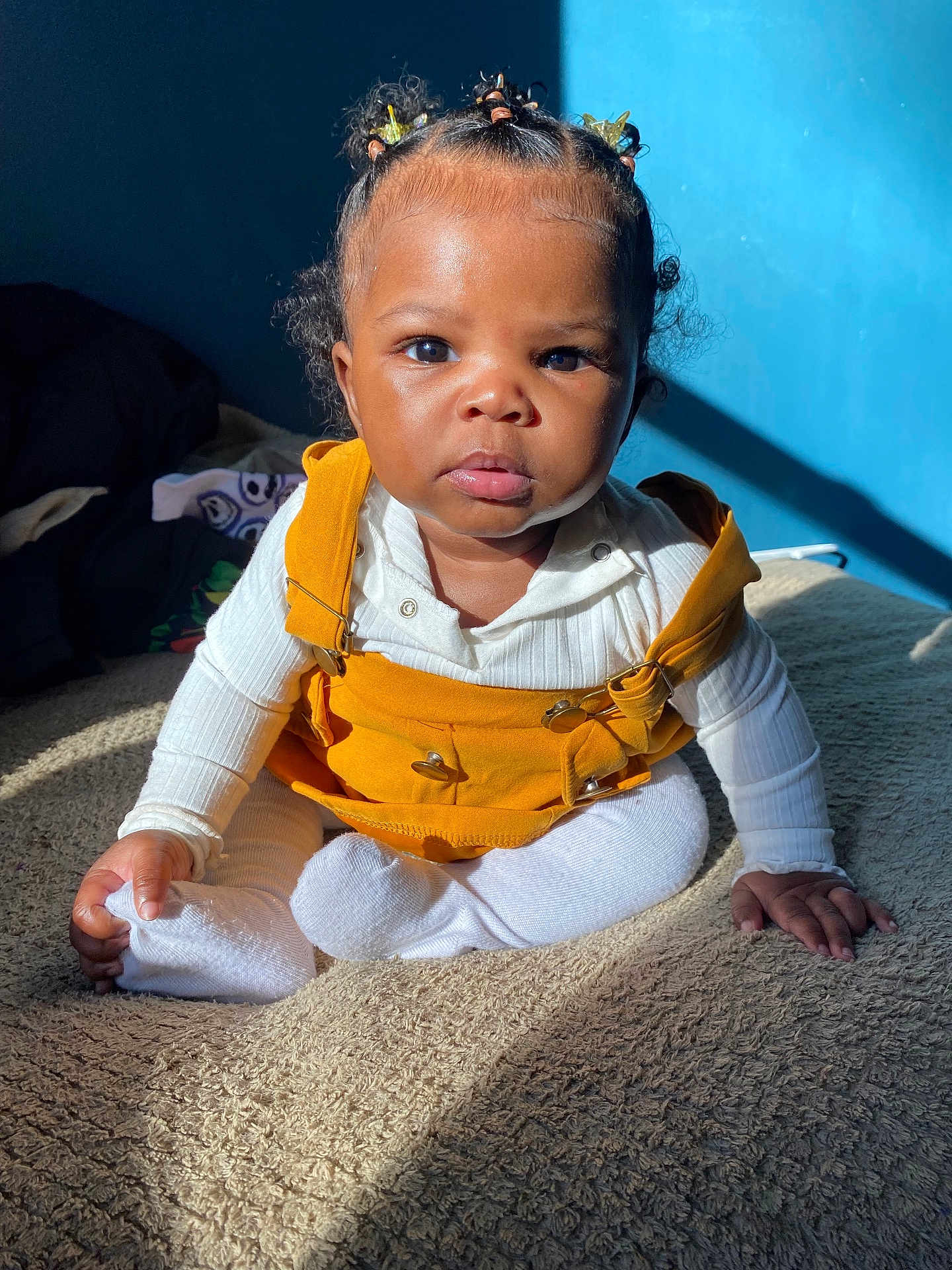 Ryleigh is registered to the contest to win money with this photo: toddler, child, baby, sitting, yellow_overalls, white_shirt, hair_accessories, curly_hair, indoors, blue_wall, blanket, sunlight, shadow, expression, portrait, cozy, clothing, person, cute, young_child