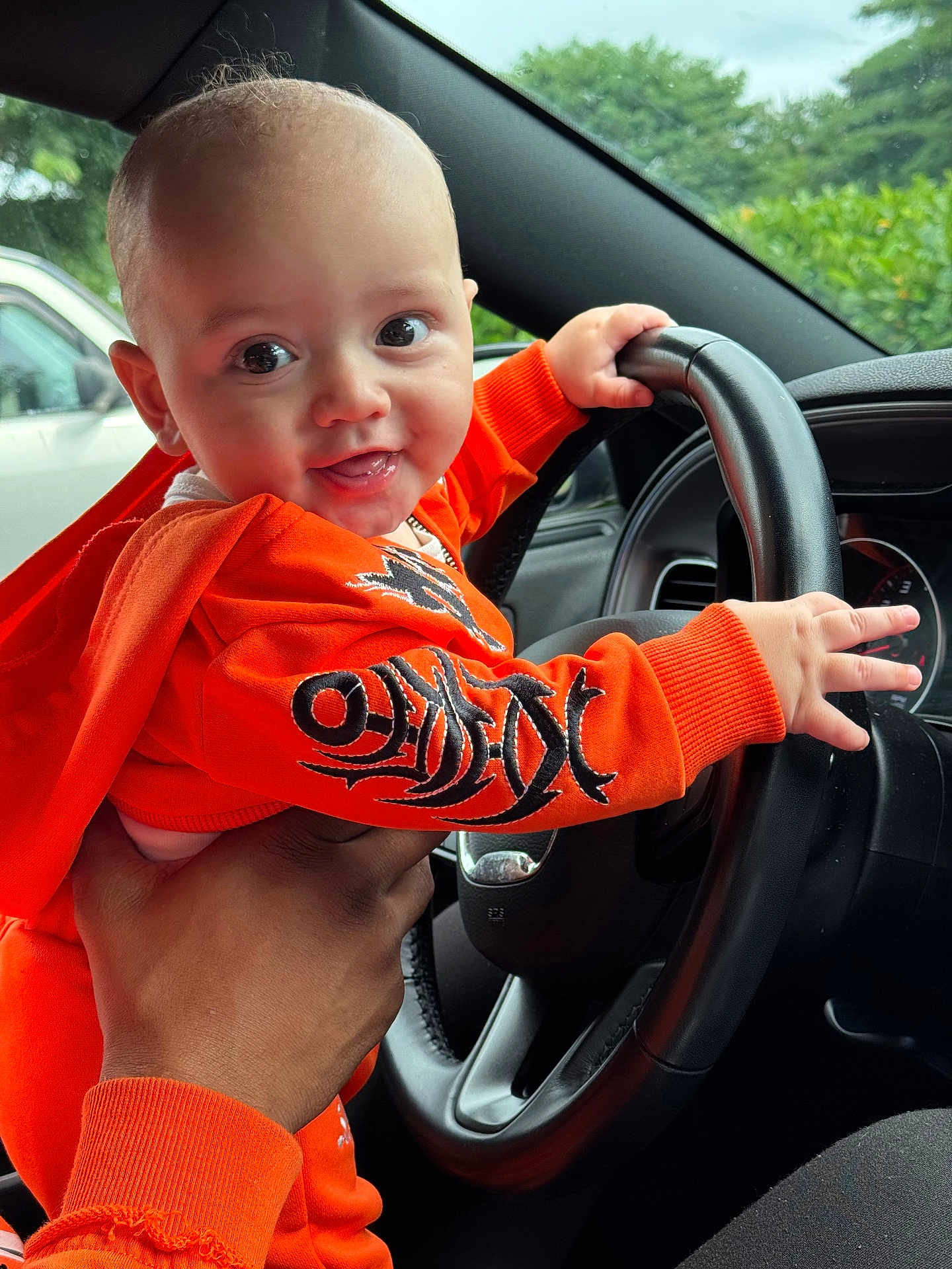 Giovanni joined the competition — help win amazing prizes! baby, child, orange_clothing, steering_wheel, car_interior, hand, smiling, cute, portrait, infant, person, window, greenery, seat, dashboard, playful, happy, closeup, casual, indoors