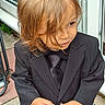 blond_hair, boy, casual_background, child, clothing, curly_hair, cute, door, expression, fashion, formal_wear, hand, outdoor, person, portrait, standing, suit, tie, window, young