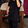 toddler, child, sunglasses, black_clothing, stylish, standing, chair, wooden_chair, beige_cushion, shoes, floor, outdoor, confident, hair, curly_hair, pose, person, young_child, casual, cool