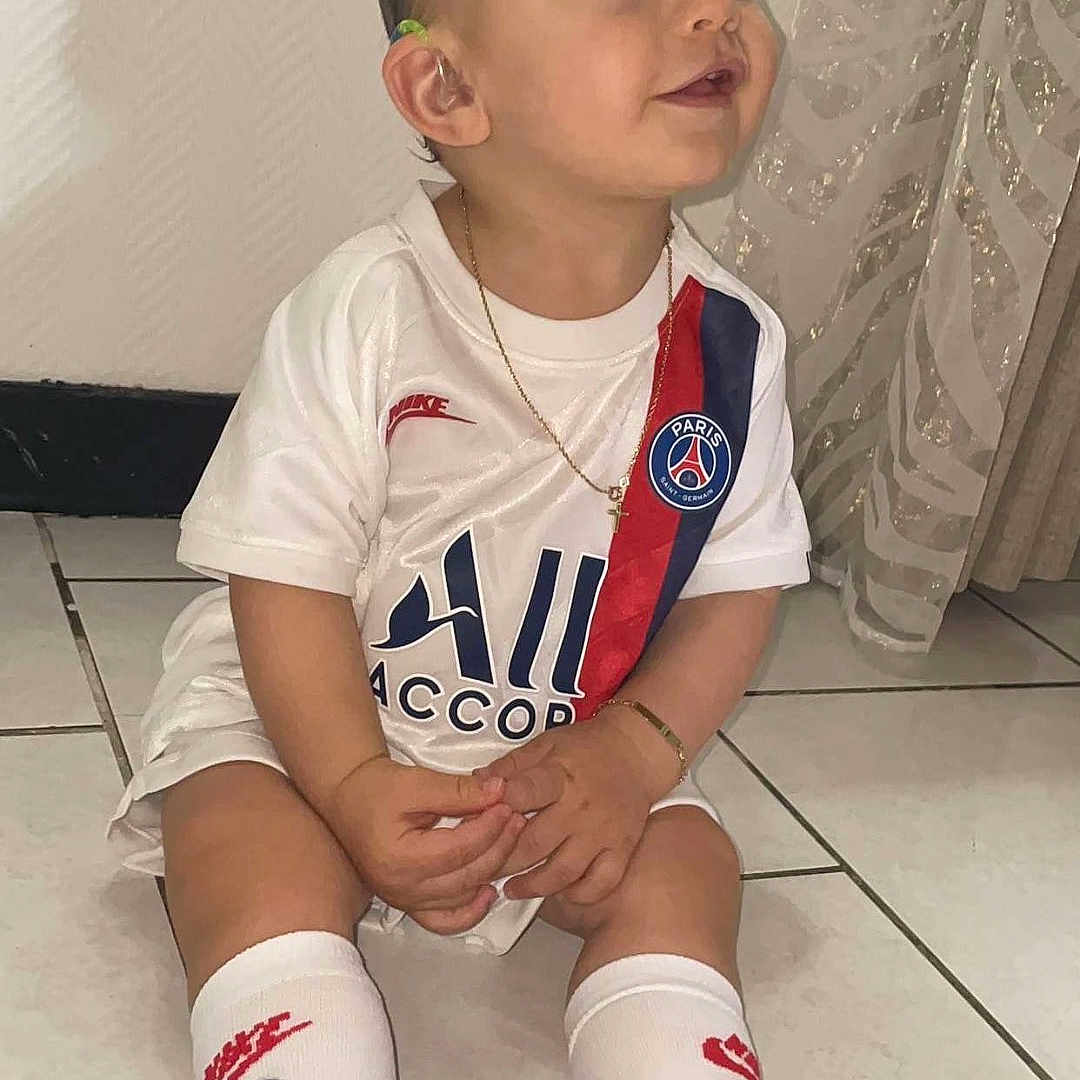 Youri participe au concours pour gagner de l'argent avec cette photo : black_sneakers, bracelet, child, curtain, cute, ear, face, floor, gold_chain, indoor, nike, paris_saint_germain, person, short_hair, sitting, smiling, soccer_jersey, tile_floor, toddler, white_socks
