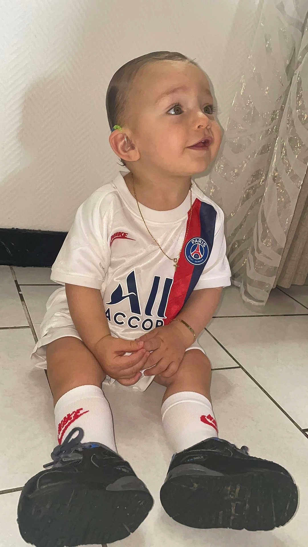 Youri participe au concours pour gagner de l'argent avec cette photo : toddler, child, sitting, soccer_jersey, paris_saint_germain, nike, white_socks, black_sneakers, floor, tile_floor, curtain, gold_chain, bracelet, indoor, smiling, cute, short_hair, ear, face, person