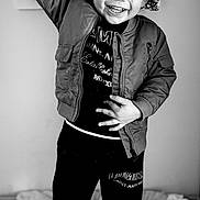 Carson is registered to the contest to win money with this photo: child, toddler, curly_hair, smiling, raising_hand, black_and_white, jacket, pants, indoor, blanket, happy, person, portrait, standing, clothing, footwear, cozy, expression, joy, casual