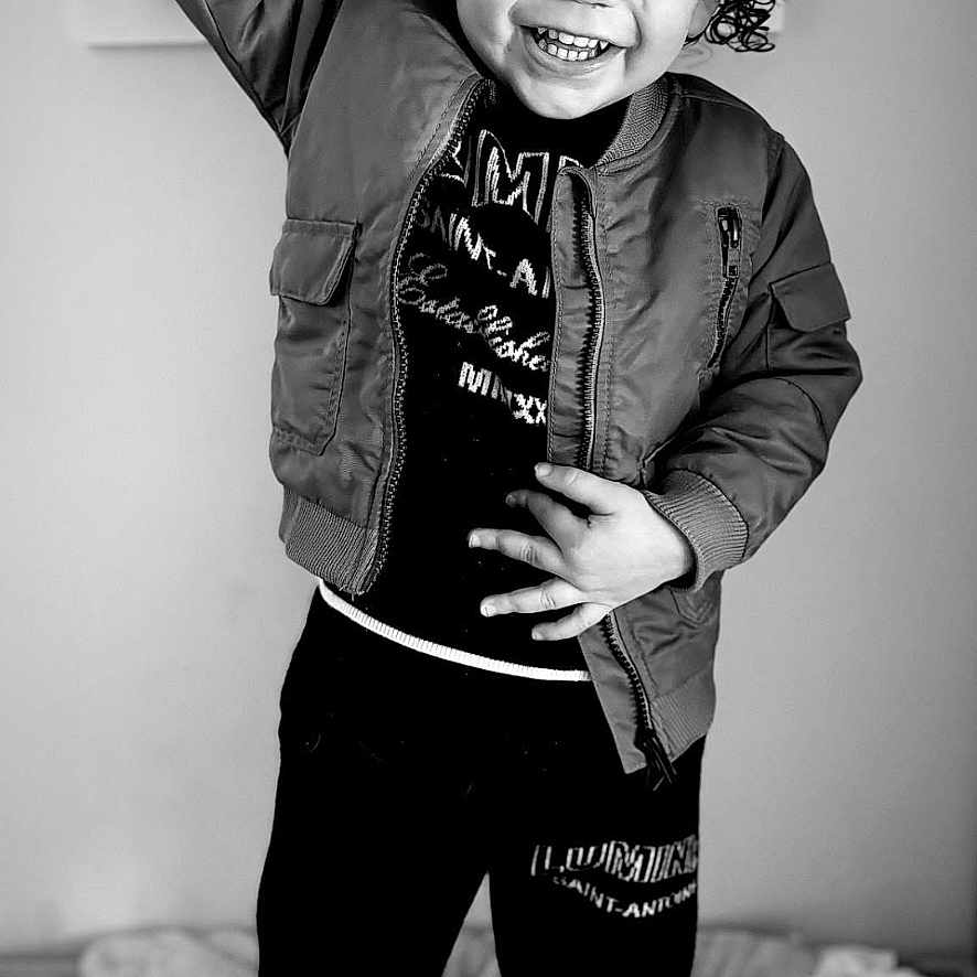 Carson is registered to the contest to win money with this photo: black_and_white, blanket, casual, child, clothing, cozy, curly_hair, expression, footwear, happy, indoor, jacket, joy, pants, person, portrait, raising_hand, smiling, standing, toddler