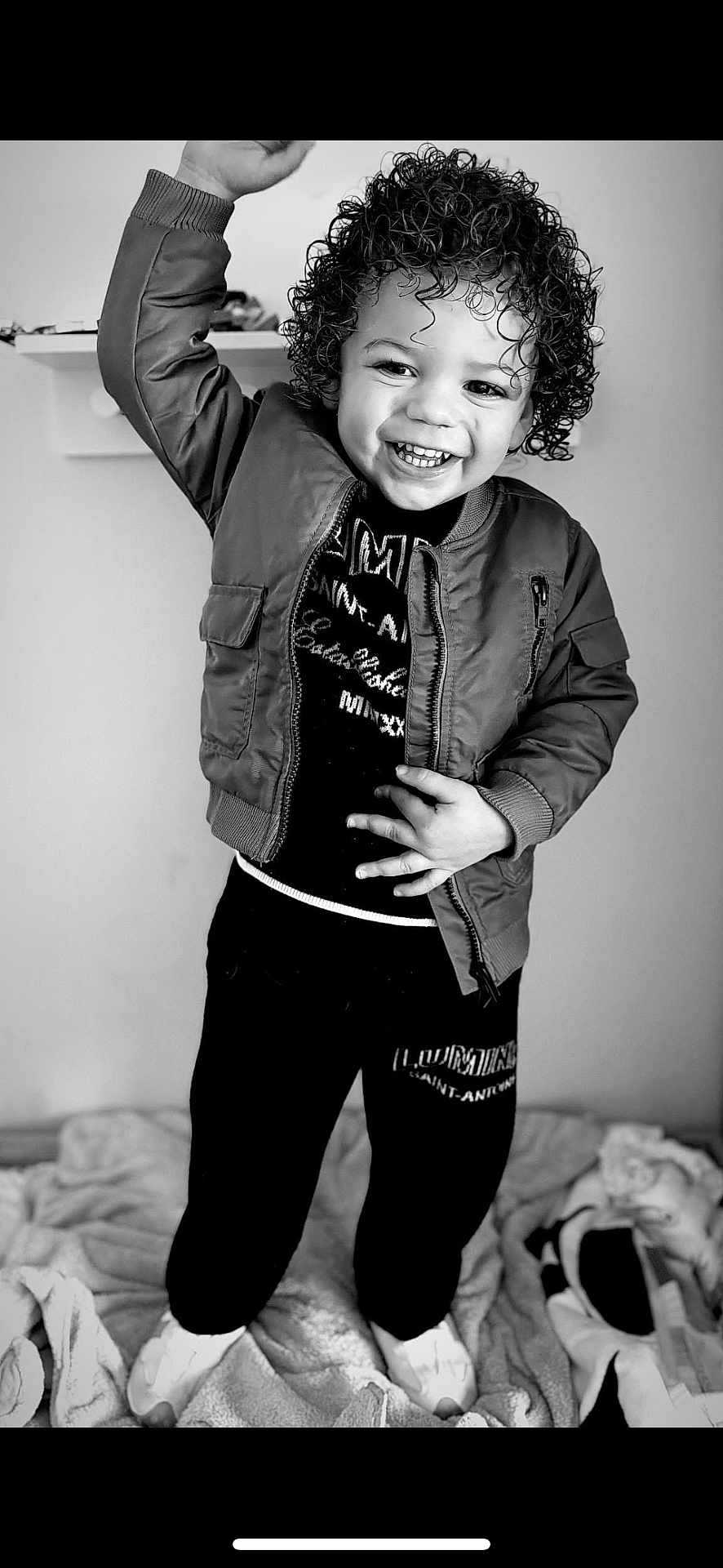 Carson is registered to the contest to win money with this photo: child, toddler, curly_hair, smiling, raising_hand, black_and_white, jacket, pants, indoor, blanket, happy, person, portrait, standing, clothing, footwear, cozy, expression, joy, casual
