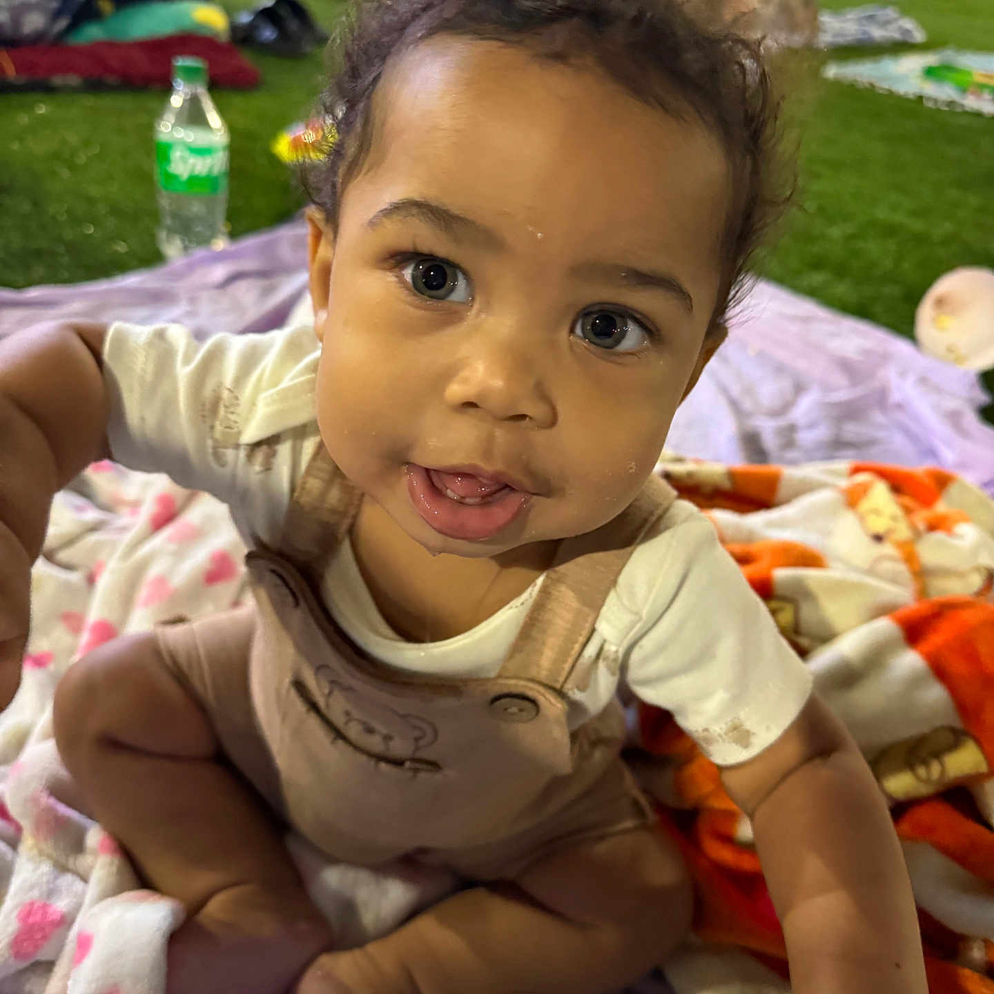 Zyaire is registered to the contest to win money with this photo: baby, child, grass, blanket, outdoor, person, curious, face, eyes, clothing, sitting, colorful, people, background, soft_lighting, infant, cute, expression, closeup, playful