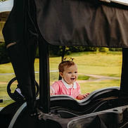 Layla is registered to the contest to win money with this photo: child, toddler, smiling, outdoor, grass, golf_cart, happy, baby, pigtails, nature, daylight, vehicle, greenery, cute, portrait, playing, pink_shirt, small_person, sunlight, joyful