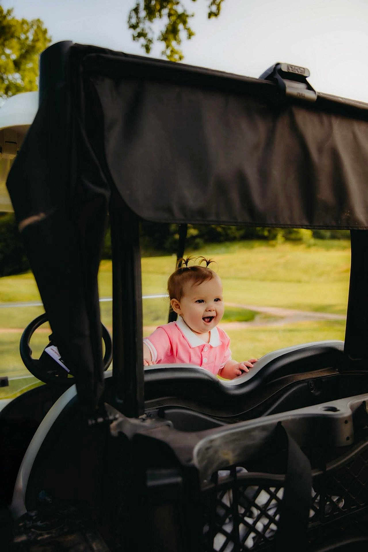 Layla is registered to the contest to win money with this photo: child, toddler, smiling, outdoor, grass, golf_cart, happy, baby, pigtails, nature, daylight, vehicle, greenery, cute, portrait, playing, pink_shirt, small_person, sunlight, joyful
