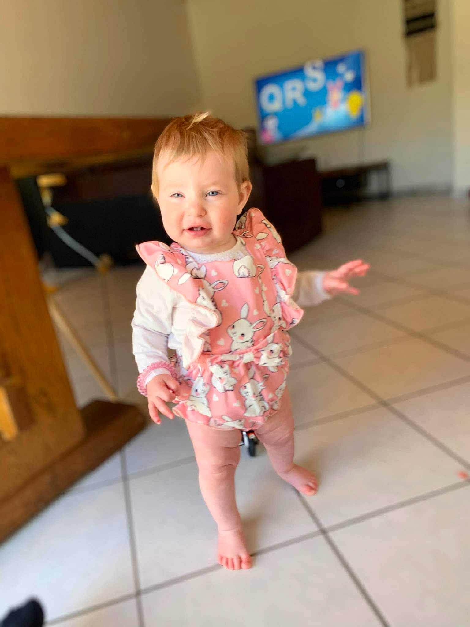 Lili participe au concours pour gagner de l'argent avec cette photo : baby, baby_toddler_clothing, barefoot, cheek, child, dress, floor, flooring, foot, fun, happy, hardwood, human_body, person, skin, sleeve, smile, television, thumb, toddler