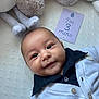 baby, blanket, blue, buttoned_clothing, cardigan, child, collared_shirt, cute, face, happy, indoors, infant, lying_down, milestone_card, plush_toys, portrait, smiling, soft_toys, text, white