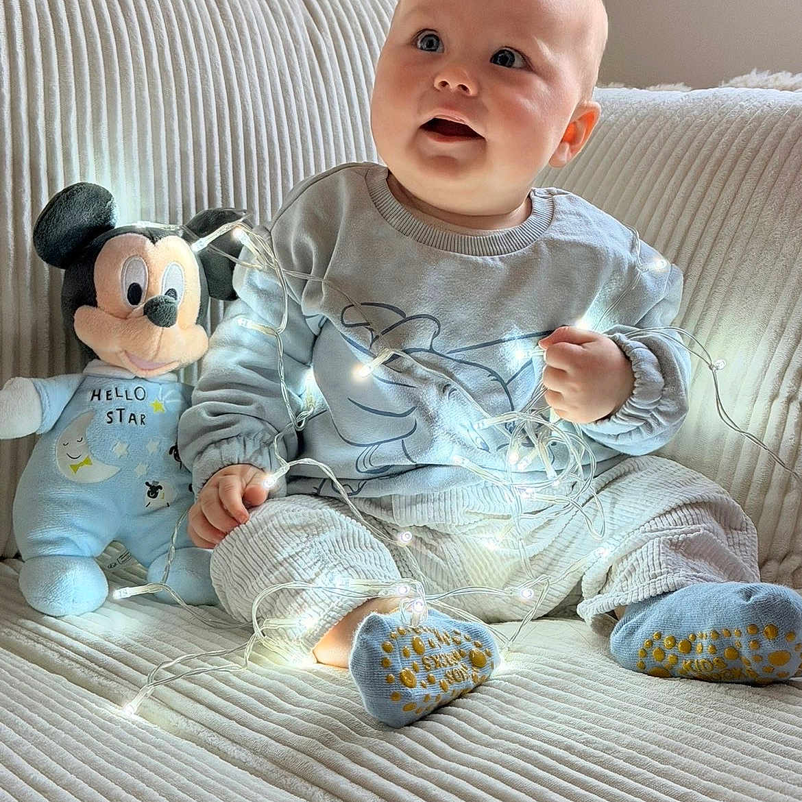 Hadriel participe au concours pour gagner de l'argent avec cette photo : baby, blue_clothing, child, couch, cozy, cute, decorative_lights, fairy_lights, happy, home, indoors, infant, mickey_mouse, plush_toy, portrait, ribbed_texture, seated, smiling, socks, soft_toys