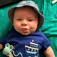 Ilyes a rejoint le concours — aidez-le/la à gagner de superbes lots ! baby, infant, child, face, tongue, hat, bucket_hat, clothing, pacifier, pacifier_clip, blue_shirt, boat_graphic, lying_down, blanket, teal_background, cute, cheeky, expression, person, indoors